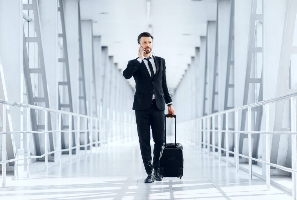 Young businessman talking on phone, walking with luggage in airport